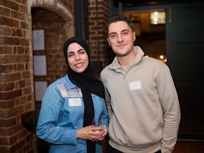 Woman in black hijab and blue denim shirt stands with man in beige pullover, both wearing name tags, smiling at camera.