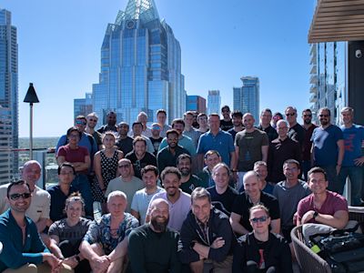 Large group photo of people on a rooftop terrace with Austin skyline and Frost Bank Tower in the background under blue sky.