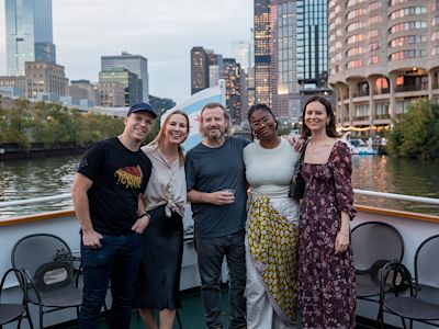 Five people posing together on a boat with city skyline and river in the background at dusk