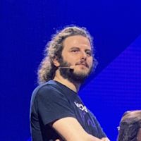 Man with long curly hair and beard wearing black t-shirt and headset microphone against bright blue background