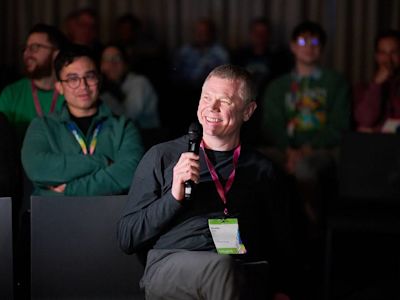 Smiling man holding microphone at conference with audience members wearing green shirts and lanyards in background