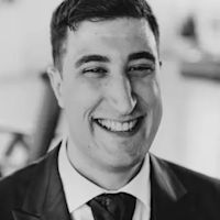 Black and white portrait of a smiling young man in a dark suit and tie with short dark hair against a blurred background.