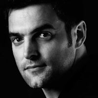 Black and white portrait of a young man with dark hair looking directly at camera against a dark background.