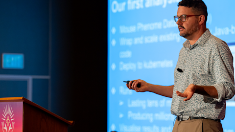 Man in glasses and patterned shirt presenting with remote control in front of blue projection screen at podium
