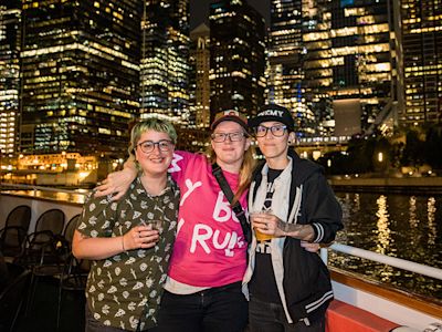 Three friends wearing glasses pose together on a boat with illuminated city skyline and waterfront at night.