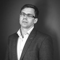 Black and white professional portrait of a man wearing glasses, a dark blazer, and white shirt against a gray background.