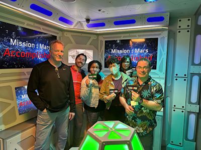 Six people posing in a futuristic space-themed room with blue lighting and Mission Mars screens, holding game devices.