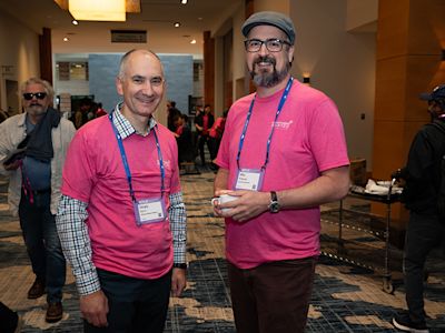 Two men wearing matching pink shirts and blue lanyards smile at a conference event in a hotel lobby.