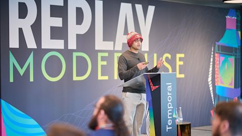 Speaker in red beanie presenting at podium with "REPLAY MODERNISE" text on large screen behind them at conference event.
