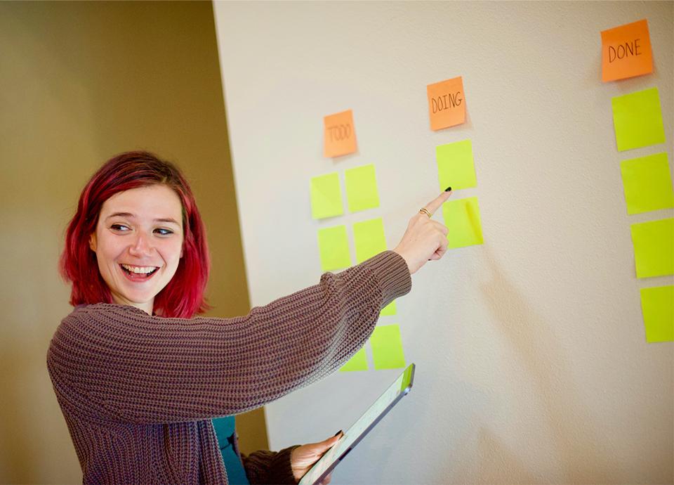 Woman with pink hair pointing to a wall of sticky notes