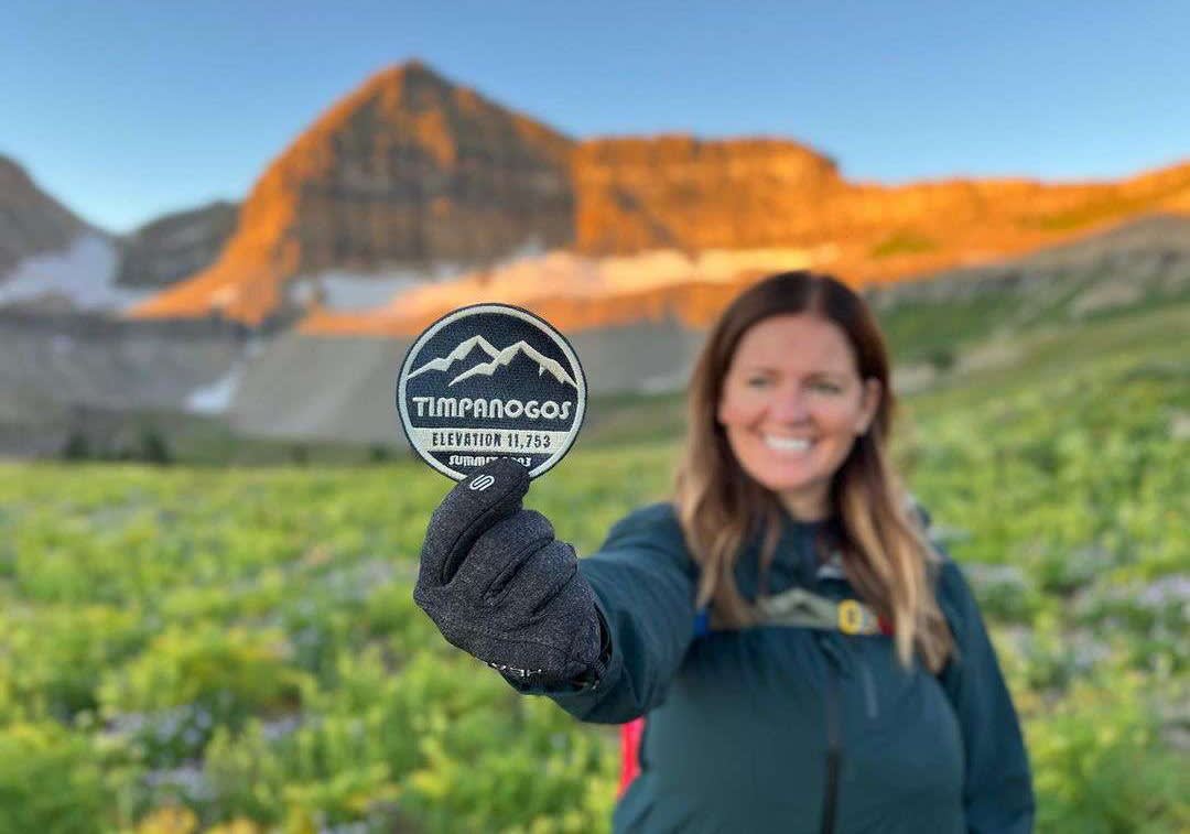 1715046544-0 - A woman holds a 2023 Timp Badge in front of Provo's Mount Timpanogos. Timpanogos Hiking Co. revived an old tradition of handing out badges to anyone who summits the mountain. 