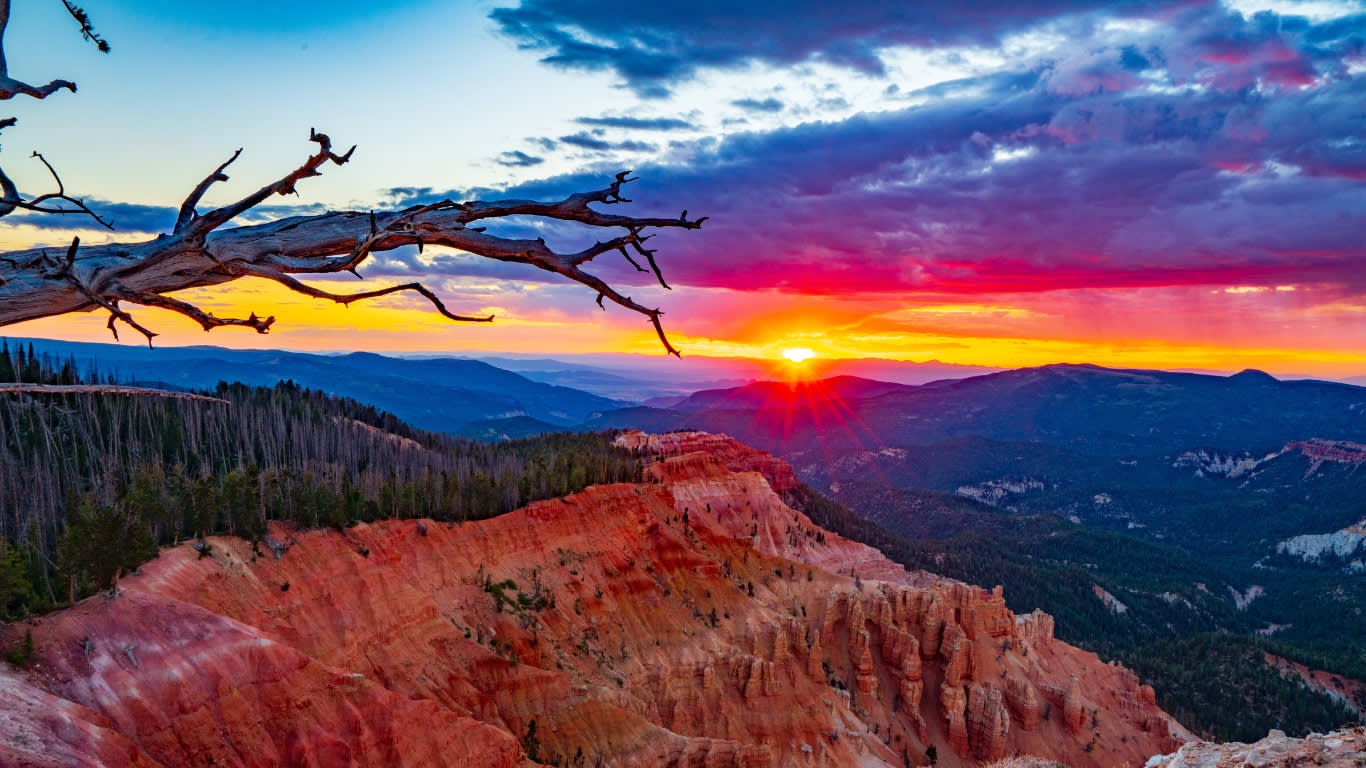 HeaderJuly - A far-off sun sets over a cliffside full red-rock spires with sprawling mountains beyond. 