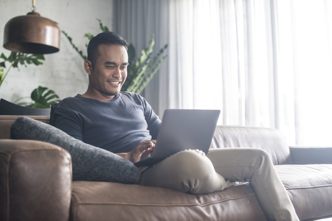 A man smiling as he works on his laptop while sitting on a couch