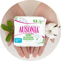 Sobre las manos de la mujer hay un paquete de compresas Ausonia. A los lados hay hojas verdes y una flor de algodón.