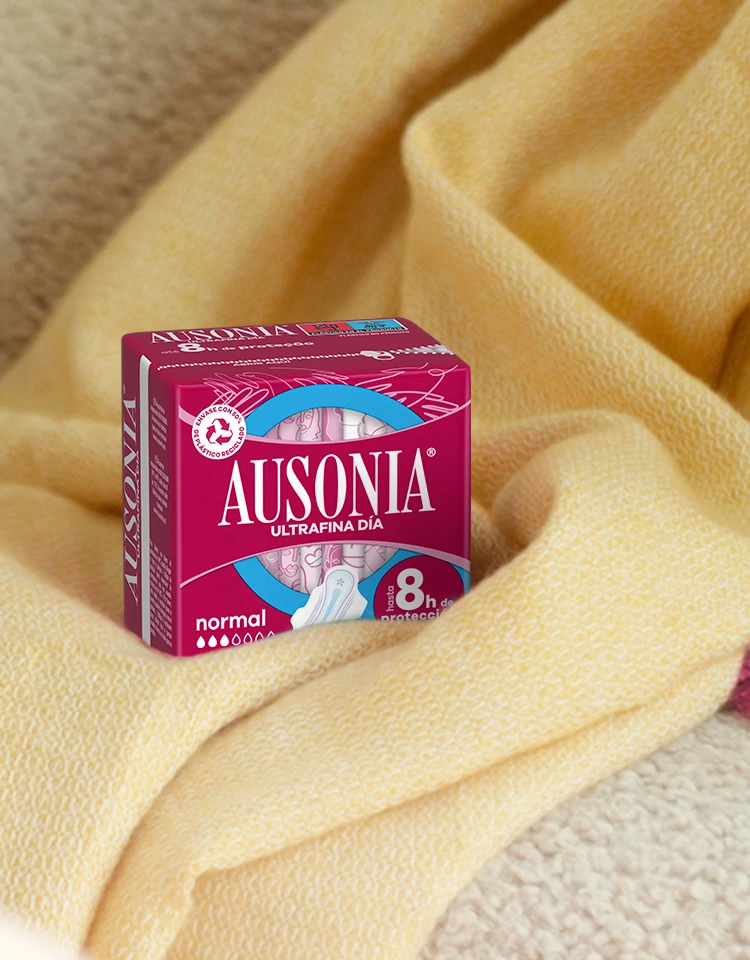 Sobre una suave manta amarilla, un paquete rosa de compresas Ausonia Ultrafina Normal con la inscripción "100% protección".