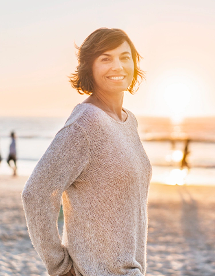 Una mujer sonriente camina por la playa. Lleva un jersey brillante. Lleva las manos en los bolsillos. 