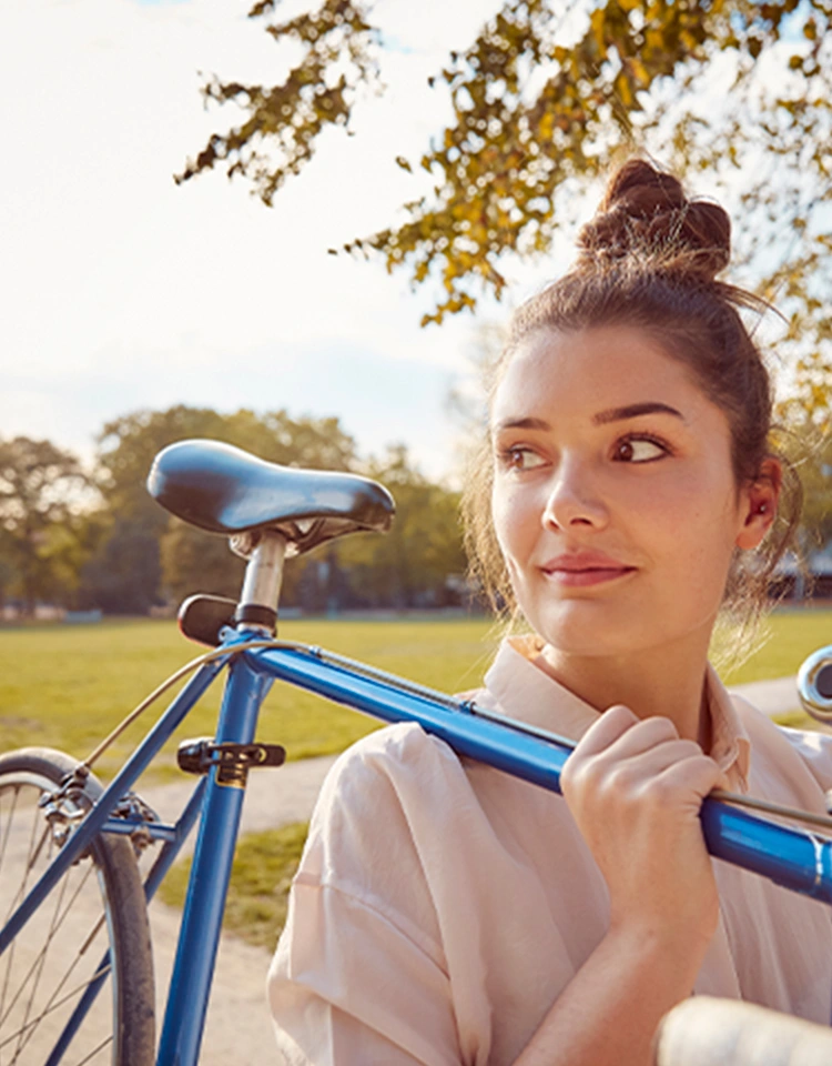 Una joven sonriente lleva una bicicleta azul apoyando su cuadro en el hombro y mira hacia un lado.