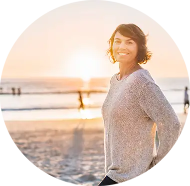 Una mujer sonriente camina por la playa. Lleva un jersey brillante. Lleva las manos en los bolsillos. 