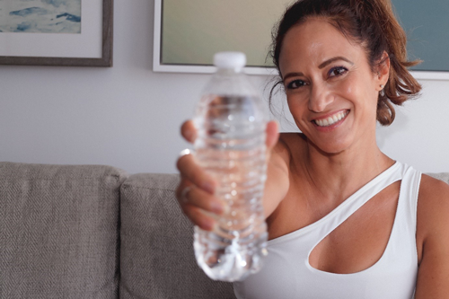 Una mujer sonriente vestida de top blanco lleva una botella de agua pequeña en la mano.