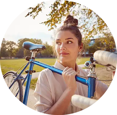 Una joven sonriente lleva una bicicleta azul apoyando su cuadro en el hombro y mira hacia un lado.