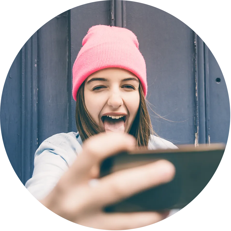 Una chica con gorra rosa sostiene un teléfono en la mano extendida. Mira la pantalla y, sonriendo, saca la lengua.
