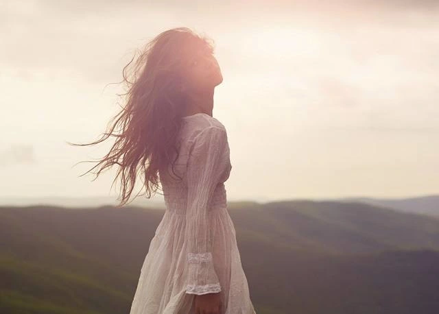 Una joven con un vestido blanco está de pie con el pelo revuelto. Tiene la cabeza inclinada hacia atrás y mira a los lados. 