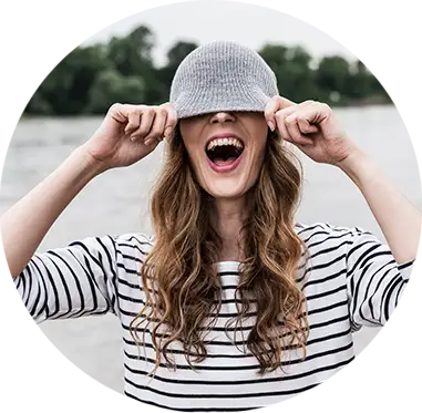 Una joven con blusa de rayas ríe y se tapa los ojos con una gorra rosa. Al fondo se ven árboles verdes.