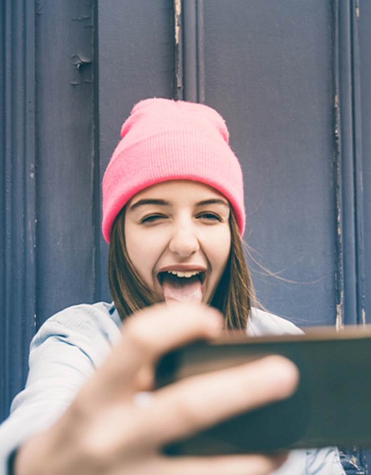 Una chica con gorra rosa sostiene un teléfono en la mano extendida. Mira la pantalla y, sonriendo, saca la lengua.