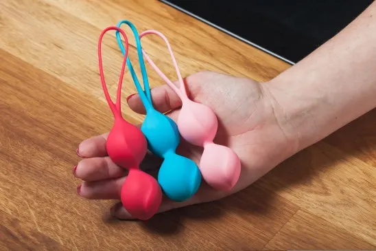 Mano de mujer sosteniendo bolas kegel rojas, azules y rosas sobre una mesa de madera