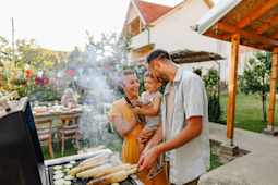 Family of three using an outdoor grill, the mother holds a child and the father looks at them while smiling.  