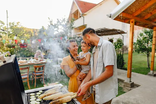 Family of three using an outdoor grill, the mother holds a child and the father looks at them while smiling.