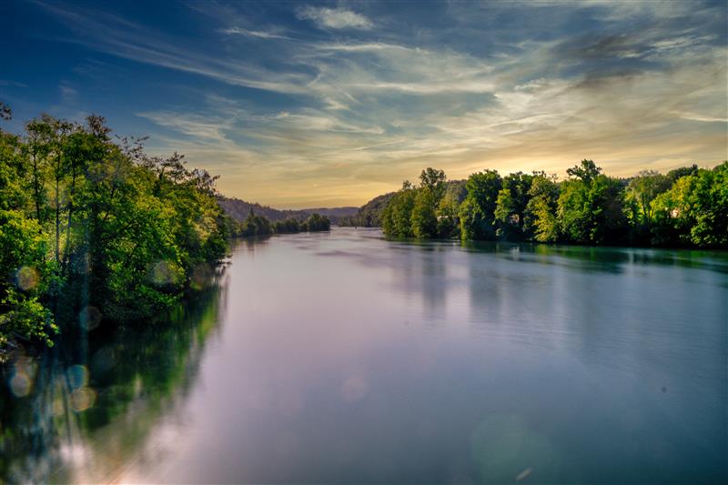 Ruhiger Fluss zwischen dichten grünen Bäumen bei Sonnenuntergang, mit weichem Licht und leicht bewölktem Himmel in einer idyllischen Naturlandschaft.