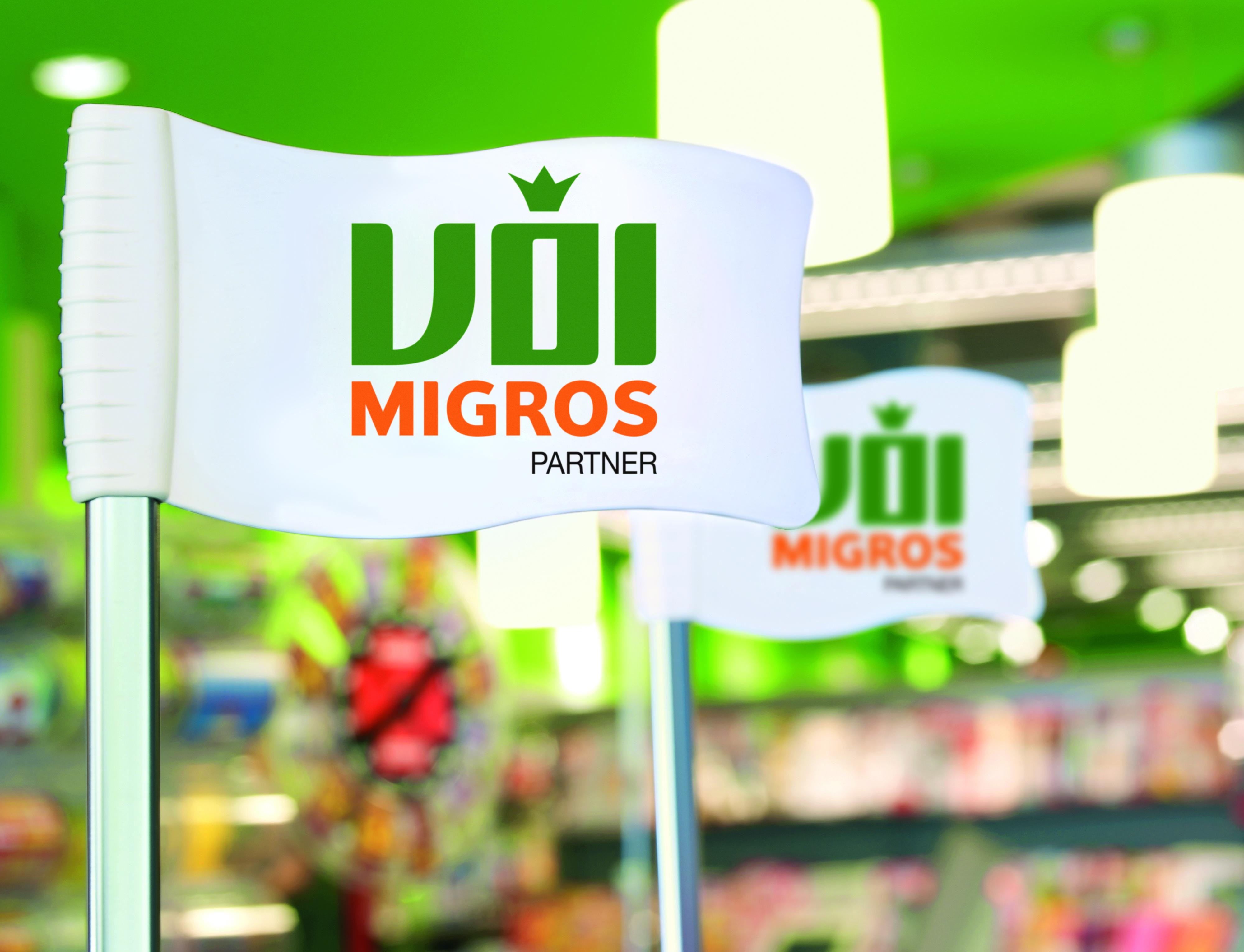 Weisse Fahne mit dem VOI Migros-Partner Logo im Vordergrund. Im Hintergrund sieht man die VOI Migros-Partner Verkaufsfläche.