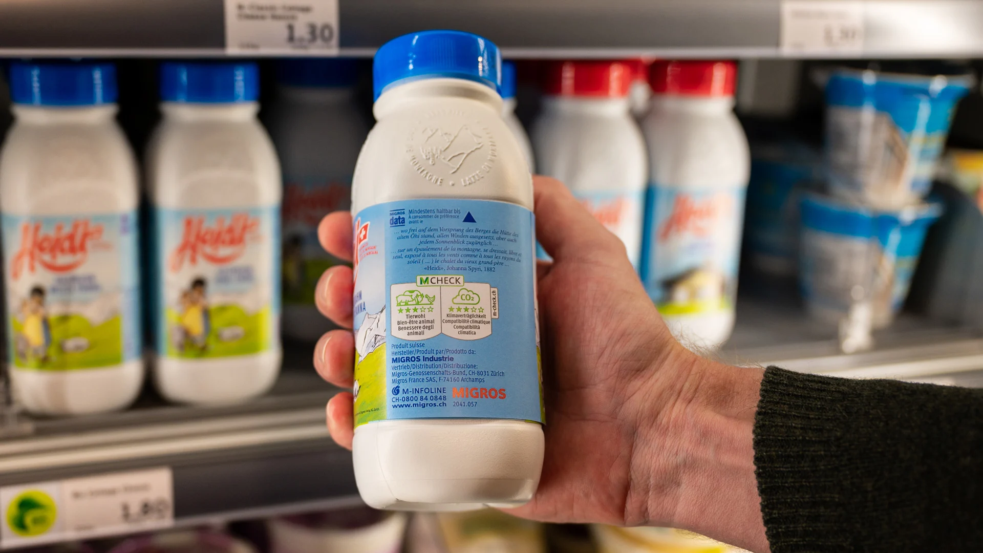 Eine Hand hält eine Milch-Flasche mit einer M-Check-Illustration.
