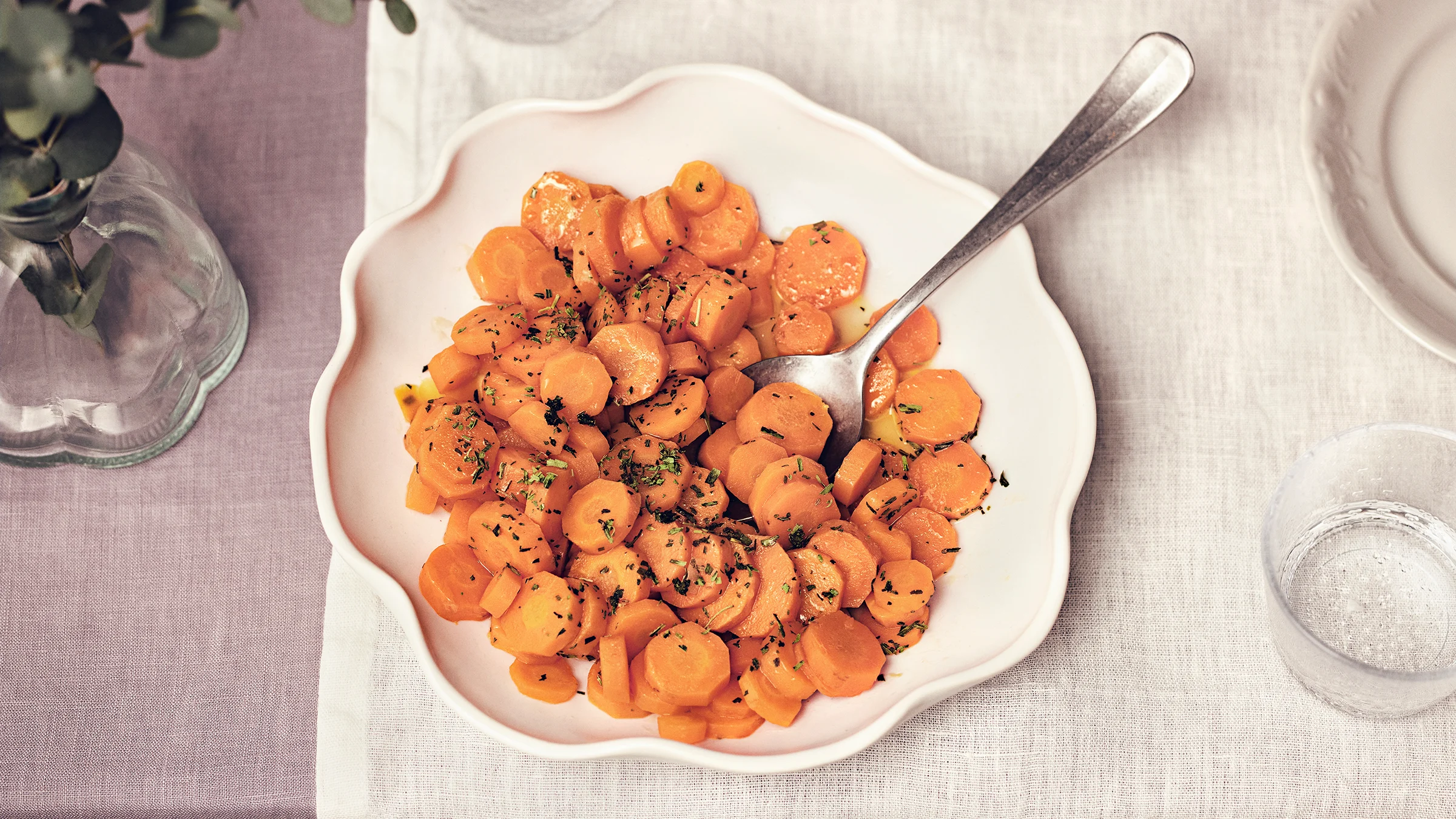 Rakuunaporkkanat vaalealla lautasella, jossa lusikka. Lautasen alla vaalea liina ja vieressä laseja, lautanen ja maljakko.