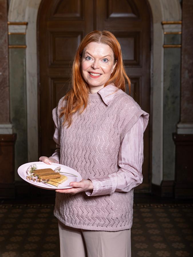 Hanne Serjo on pukeutunut violettiin kauluspaitaan sekä violettiin liivineuleeseen. Hänellä on käsissään valkoinen lautanen, jolle on aseteltu kolme taikinapatukkaa.