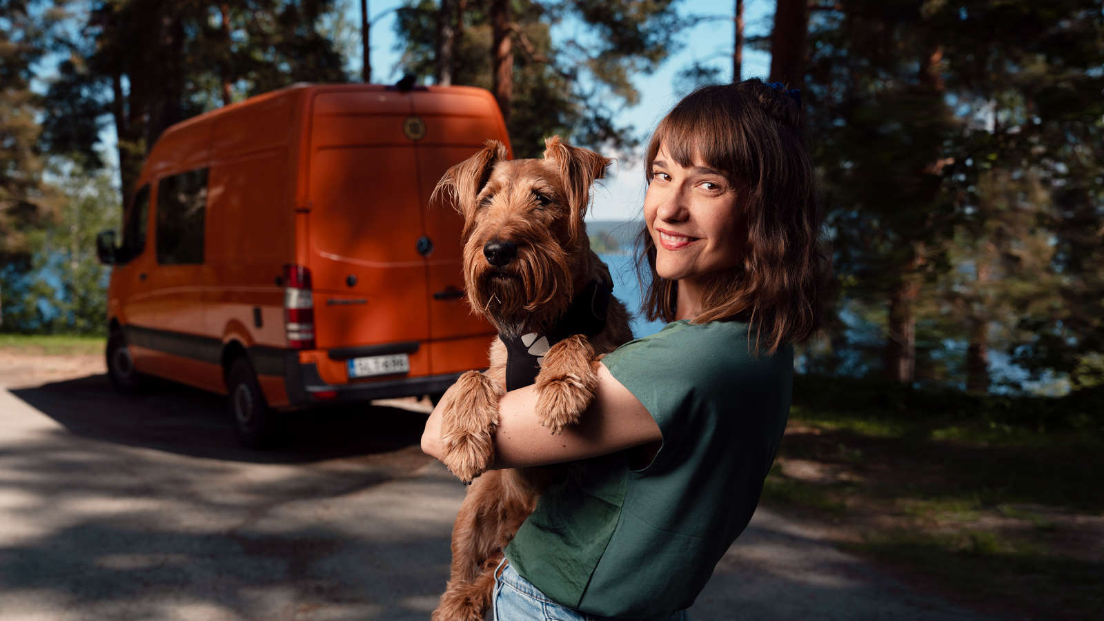 Nainen pitää sylissään koiraa, taustalla näkyy pakettiauto ja puiden lomasta pilkottava järvi