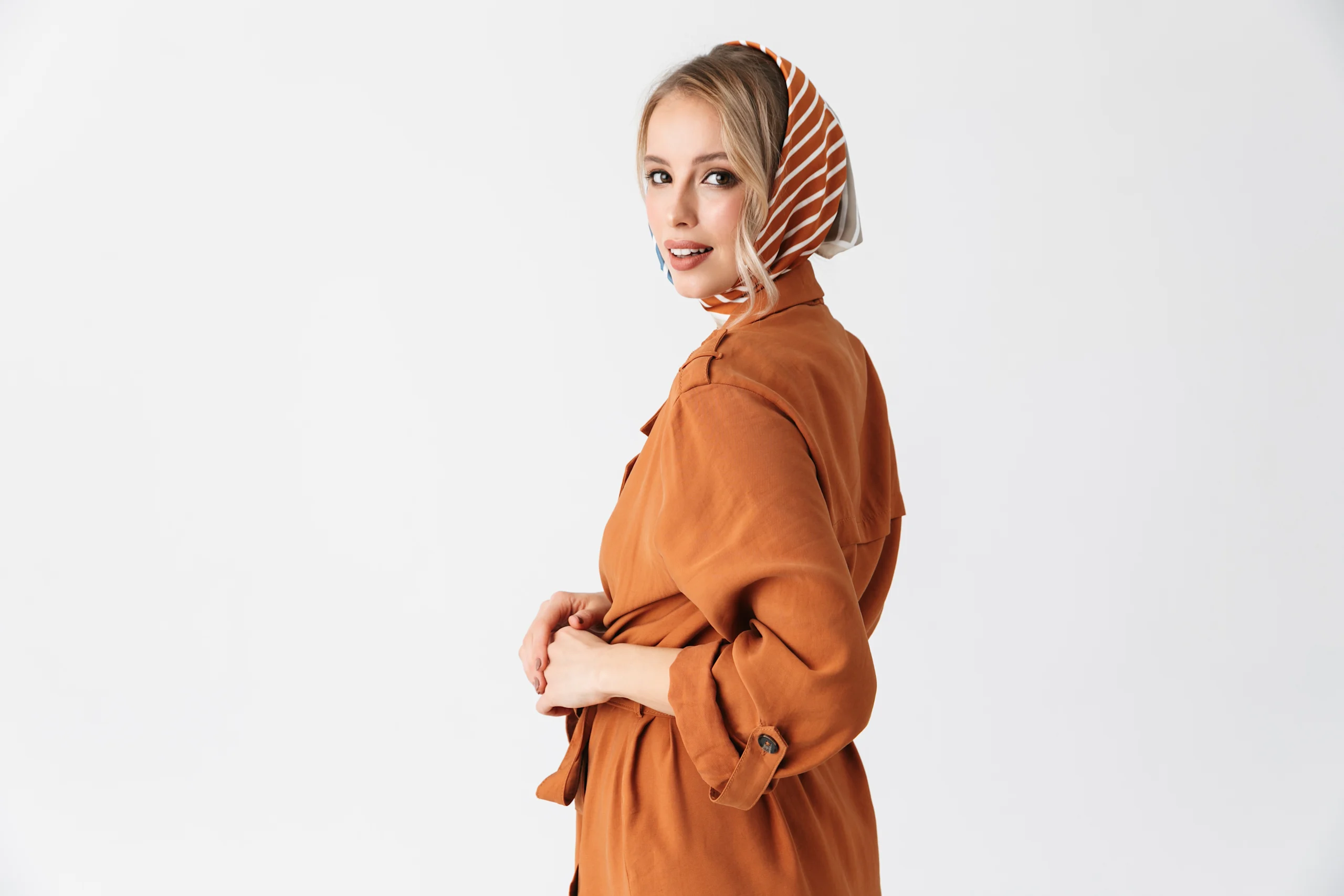 Murretun oranssiin trenssiin pukeutunut nainen katsoo kameraan. Päässä tyylikäs silkkihuivi.