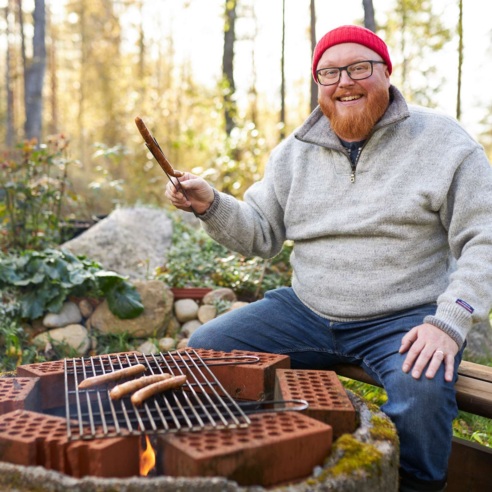 Roy Norro istuu nuotiogrillin ääressä. Grillissä on ritilän päällä makkaroita.