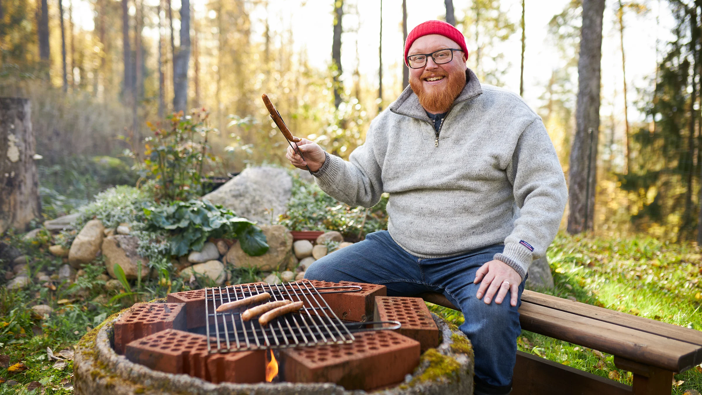 Roy Norro istuu nuotiogrillin ääressä. Grillissä on ritilän päällä makkaroita.