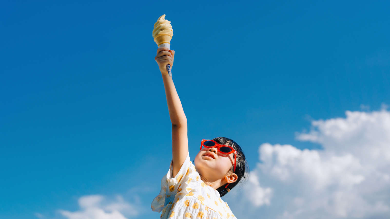 Lapsi joka pitää kädessään jäätelötuuttia sinistä taivasta vasten.