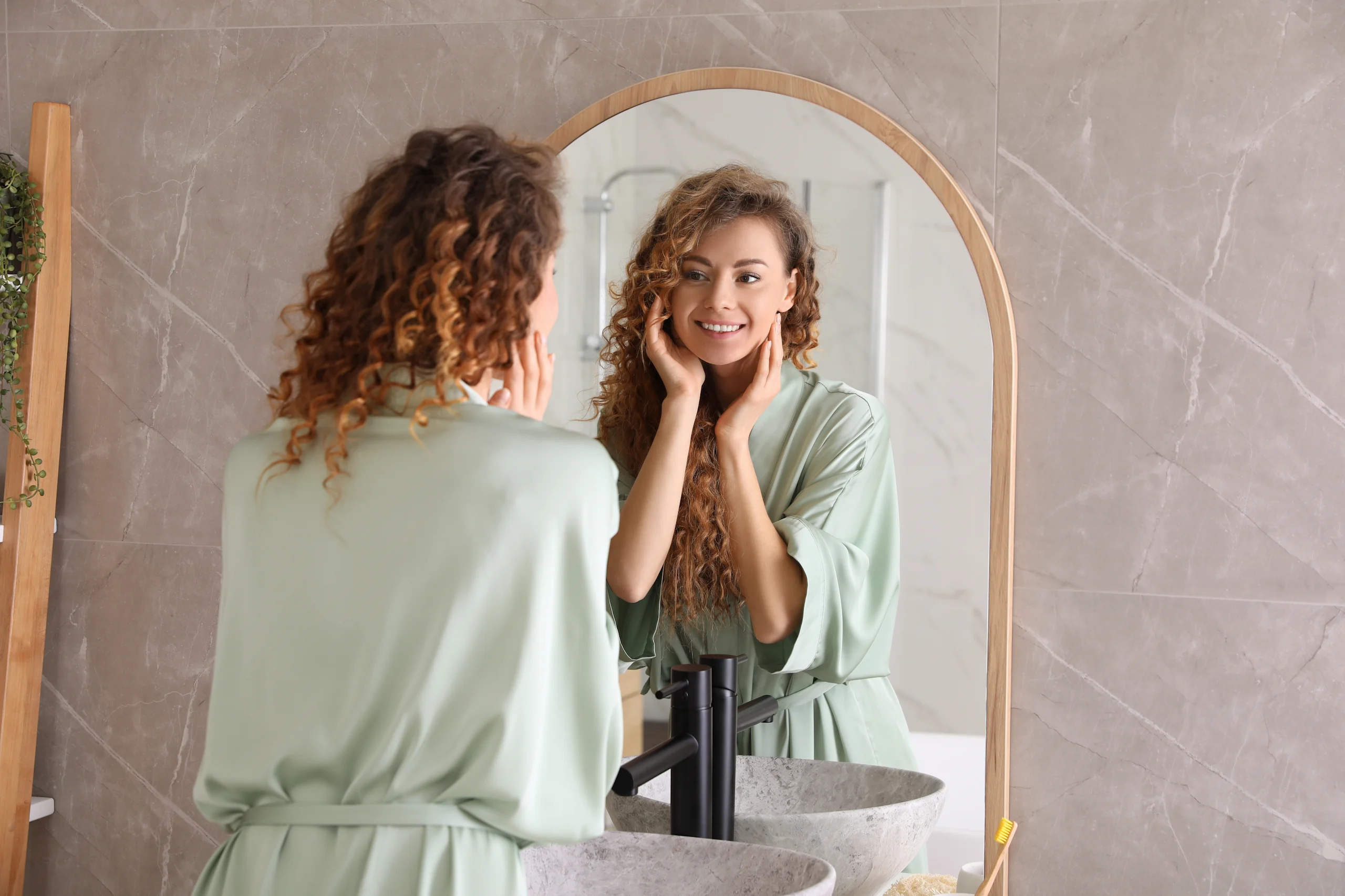 Kiharahiuksinen nainen katsoo itseään kylpyhuoneen peilistä