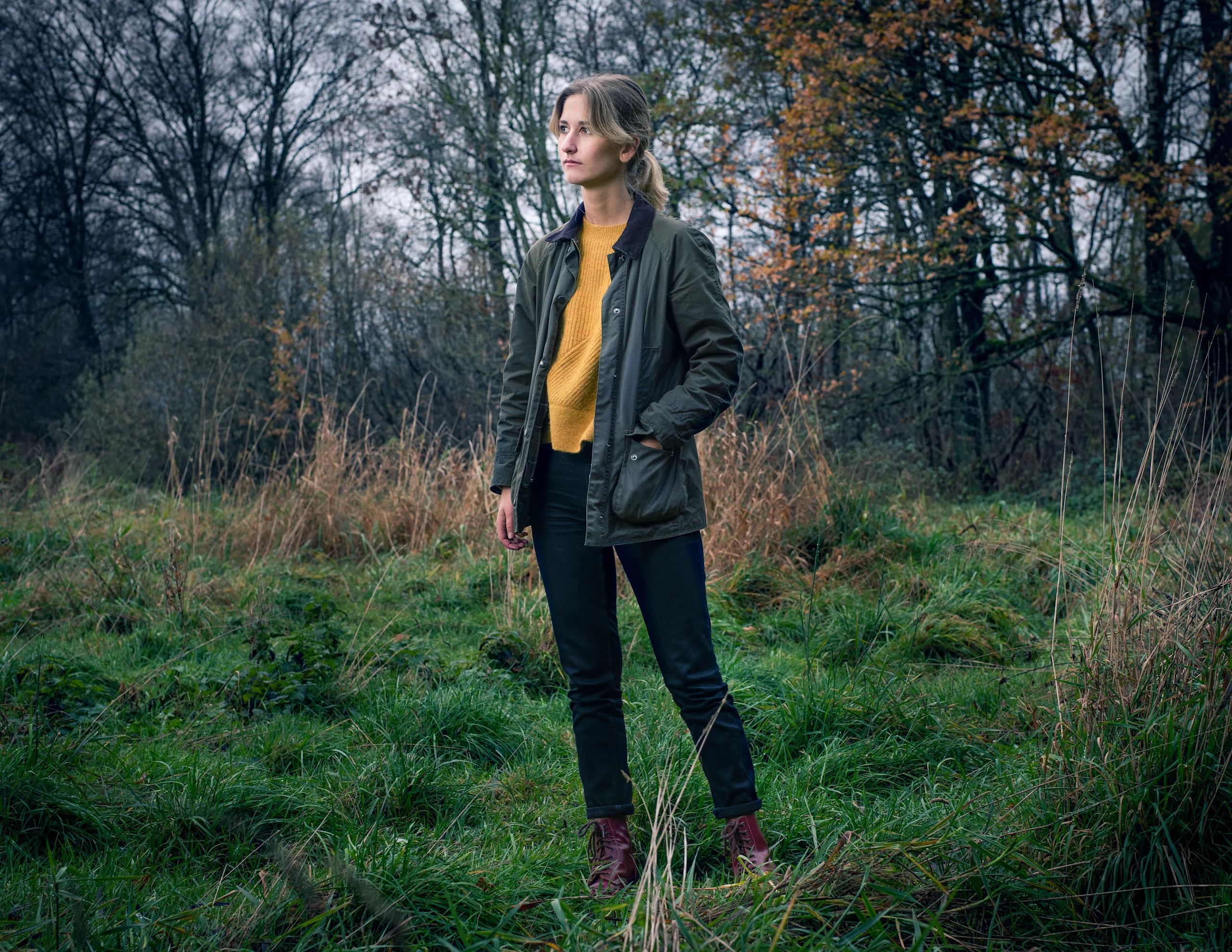 Tummanvihreään navettatakkiin ja sinapinkeltaiseen paitaan pukeutunut nainen metsässä