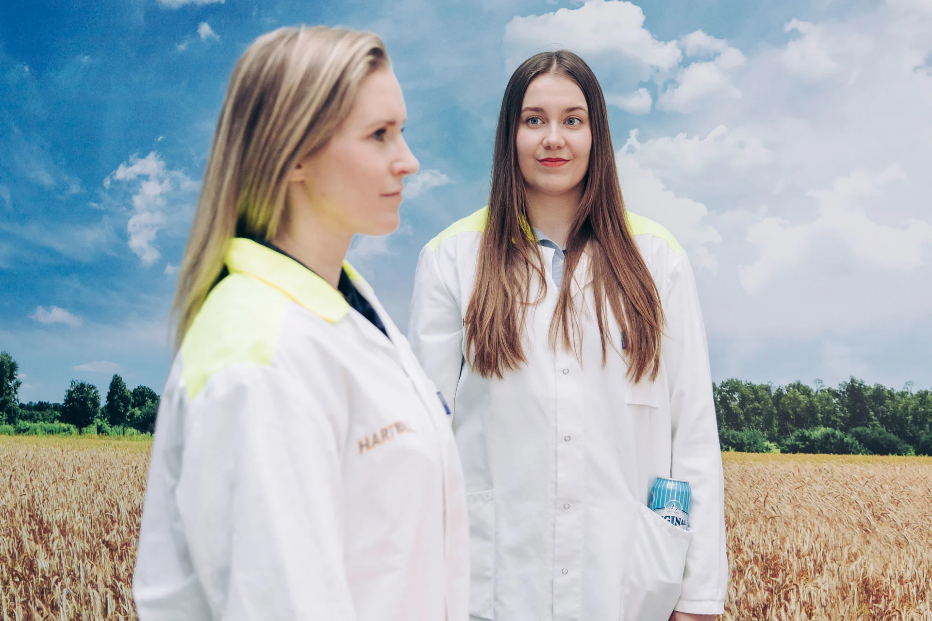 Kaksi naista seisoo peltokuvan edessä laboratoriotakeissa, toisen taskussa näkyy Long Drink -juomatölkki.