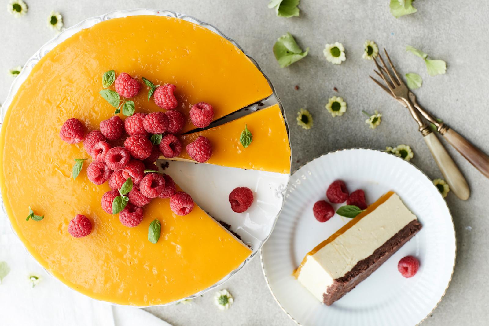 Mangojuustokakku, joka koristelu vadelmilla ja mintunlehdillä. Kakusta leikattu yksi pala kakkuvadille ja yksi lautaselle. Lautasen vieressä kaksi leivoshaarukkaa.