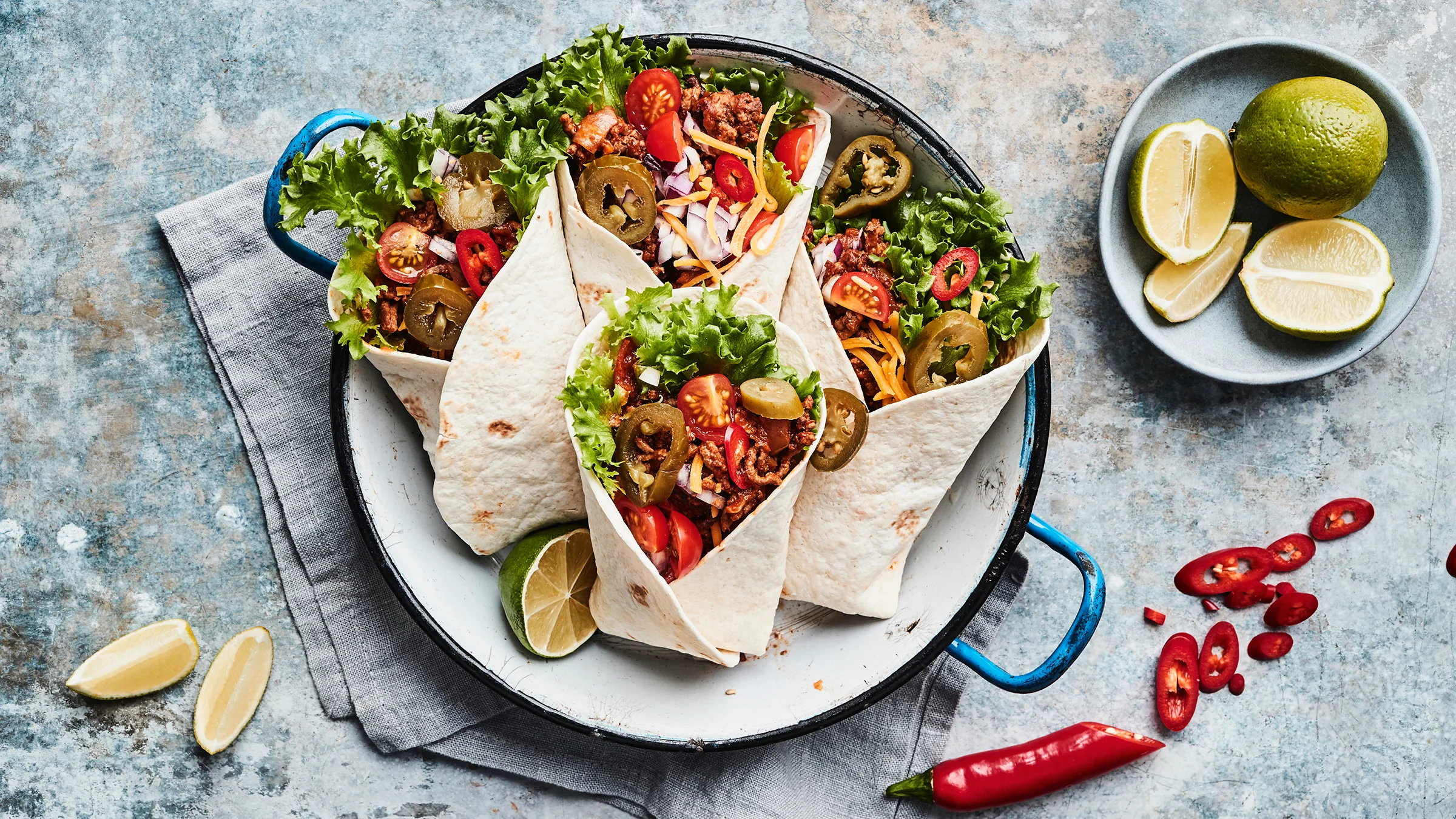 Jauhelihalla ja kasviksilla täytettyjä tortilloja tarjoilulautasella. Lautasen vieressö limelohkoja ja tuoretta chiliä pilkottuna.