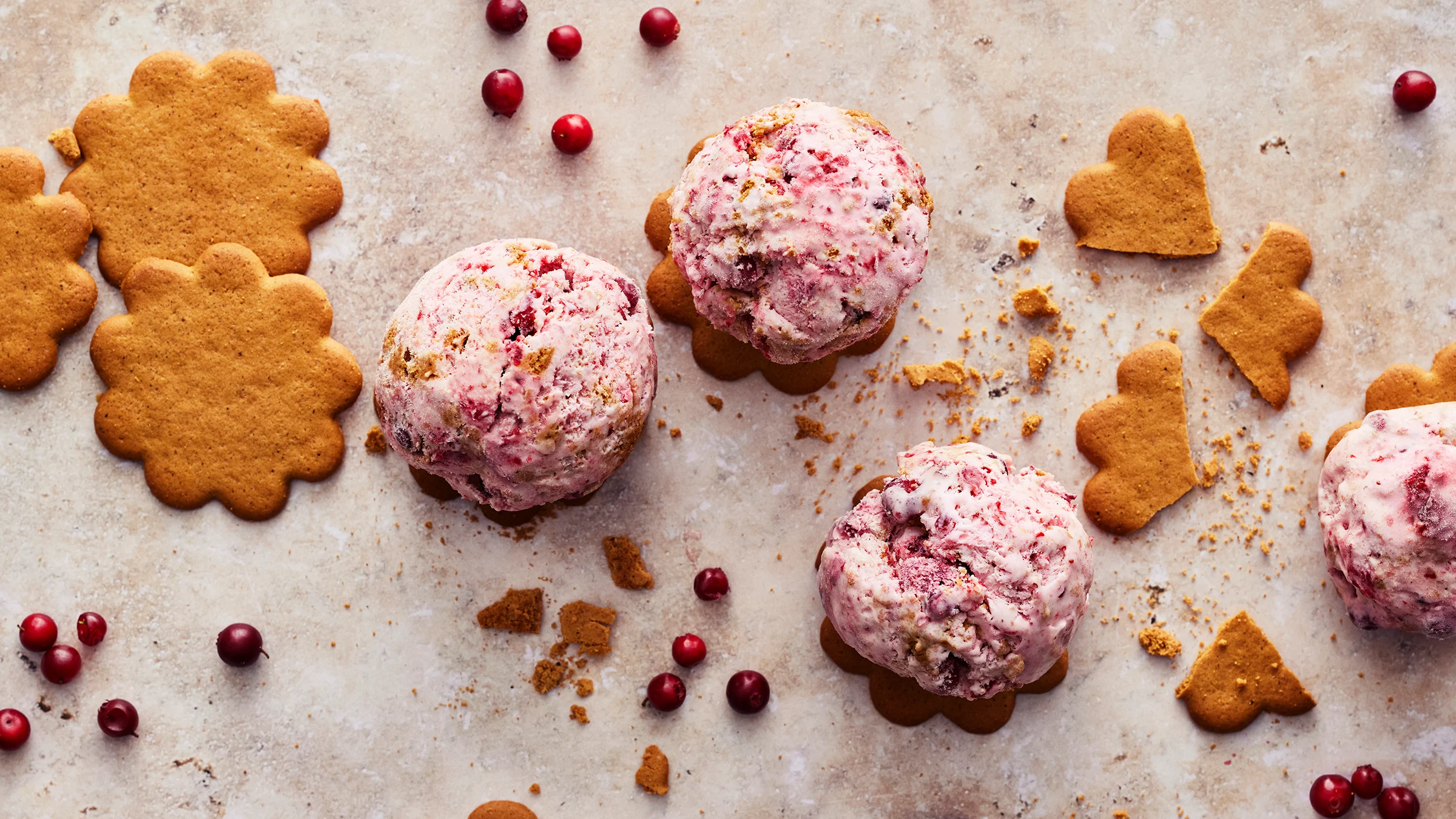 Neljä palloa piparkakku-puolukkajäätelöä piparkakkuje päällä. Pöytätasolla kokonaisia ja murennettuja piparkakkuja ja puolukoita.