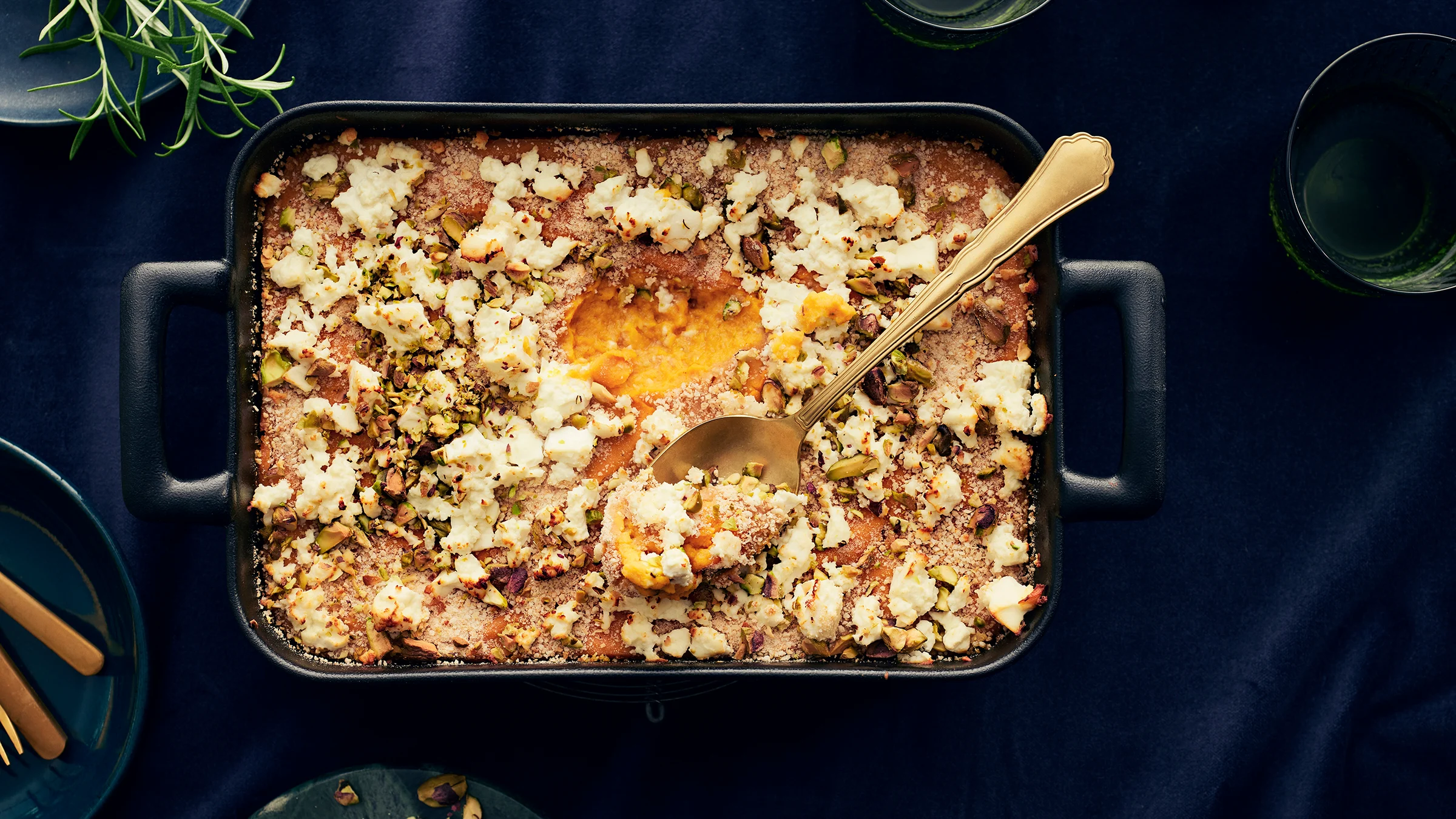 Feta-pistaasikuorrutteinen bataattilaatikko tummassa uunivuoassa. Vuoassa lusikka, jolla laatikkoa on kauhaistu.