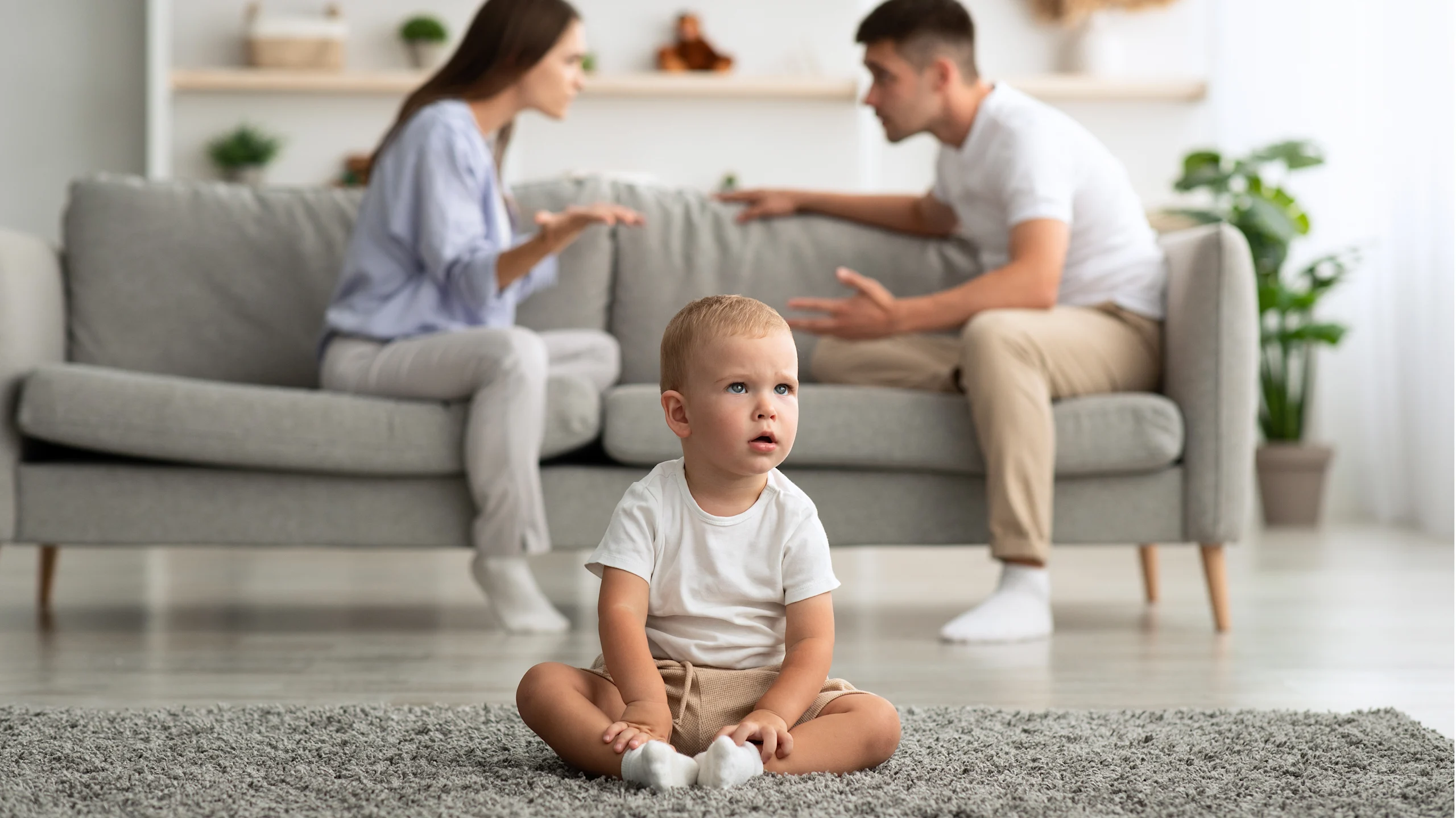 Vauva istuu etualalla, takana sohvalla riitelevät vanhemmat