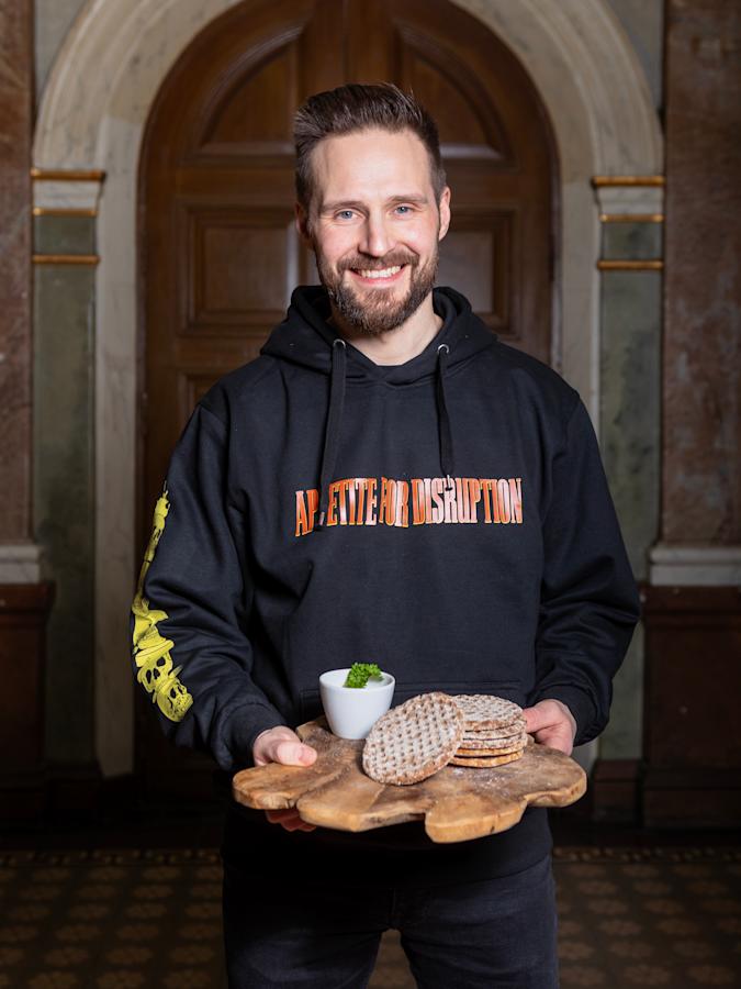 Teppo Ylä-Hemmilän mustassa paidassa lukee ”Apetite for disruption”. Käsissään hän pitelee puista tarjotinta, jolle on pinottu kuusi kappaletta pyöreiltä näkkileiviltä näyttäviä leipiä.